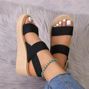 Sandali donna con zeppa in sughero, cinturini neri, indossati su tappeto grigio. Accessorio moda estivo, comodi e trendy.