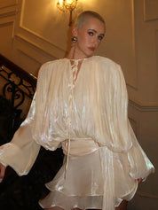 Donna con abito elegante in tessuto lucido color crema, scollatura a laccio, maniche lunghe, in posa su scala interna. Moda serale, stile glamour.
