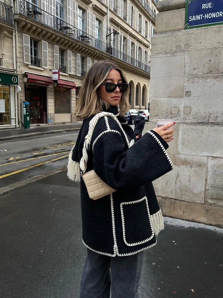 Donna con cappotto nero con dettagli bianchi, occhiali da sole e borsa beige, cammina in strada parigina. Moda autunnale, stile urbano chic.