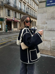 Donna con cappotto nero con dettagli bianchi, occhiali da sole e borsa beige, cammina in strada parigina. Moda autunnale, stile urbano chic.
