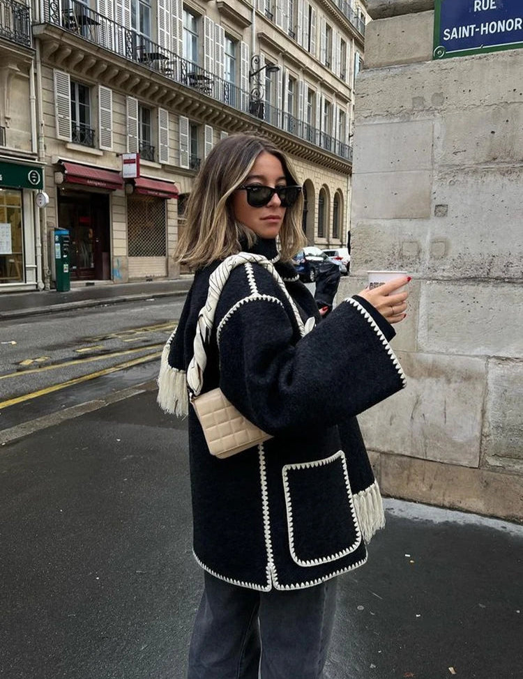 Donna con cappotto nero con dettagli bianchi, occhiali da sole e borsa beige, cammina in strada parigina. Moda autunnale, stile urbano chic.