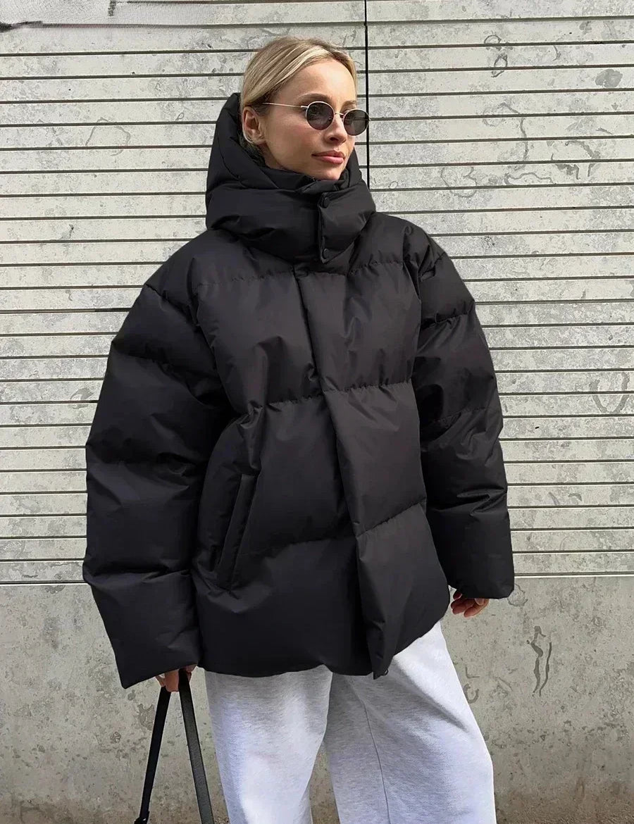 Giacca invernale oversize nera, donna con occhiali da sole, pantaloni grigi, sfondo urbano. Moda streetwear, cappotto imbottito caldo.