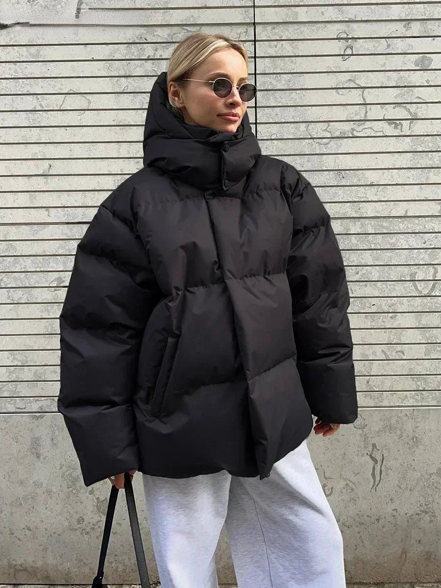 Giacca invernale oversize nera, donna con occhiali da sole, pantaloni grigi, sfondo urbano. Moda streetwear, cappotto imbottito caldo.