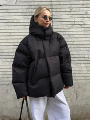 Giacca invernale oversize nera, donna con occhiali da sole, pantaloni grigi, sfondo urbano. Moda streetwear, cappotto imbottito caldo.