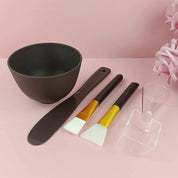 Set ciotola e strumenti per maschera viso su sfondo rosa. Include ciotola nera, spatola, pennelli e misurino. Accessori skincare essenziali.