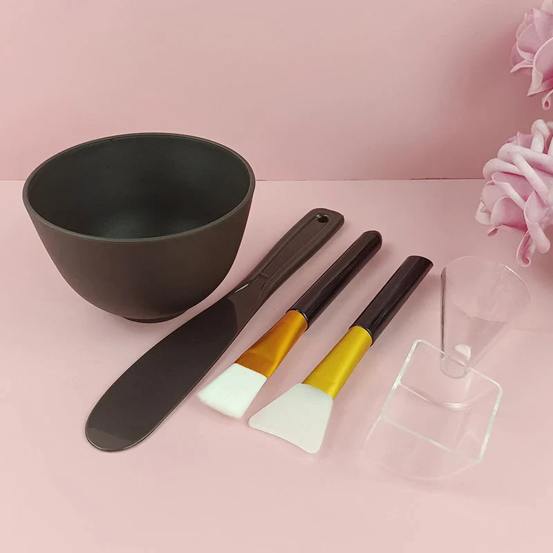 Set ciotola e strumenti per maschera viso su sfondo rosa. Include ciotola nera, spatola, pennelli e misurino. Accessori skincare essenziali.