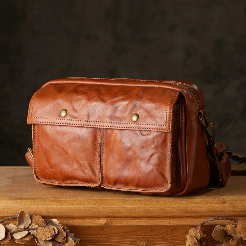 Borsa a tracolla in pelle marrone vintage su tavolo di legno, design con doppia tasca frontale e chiusura a bottone, ideale per uomo e donna.