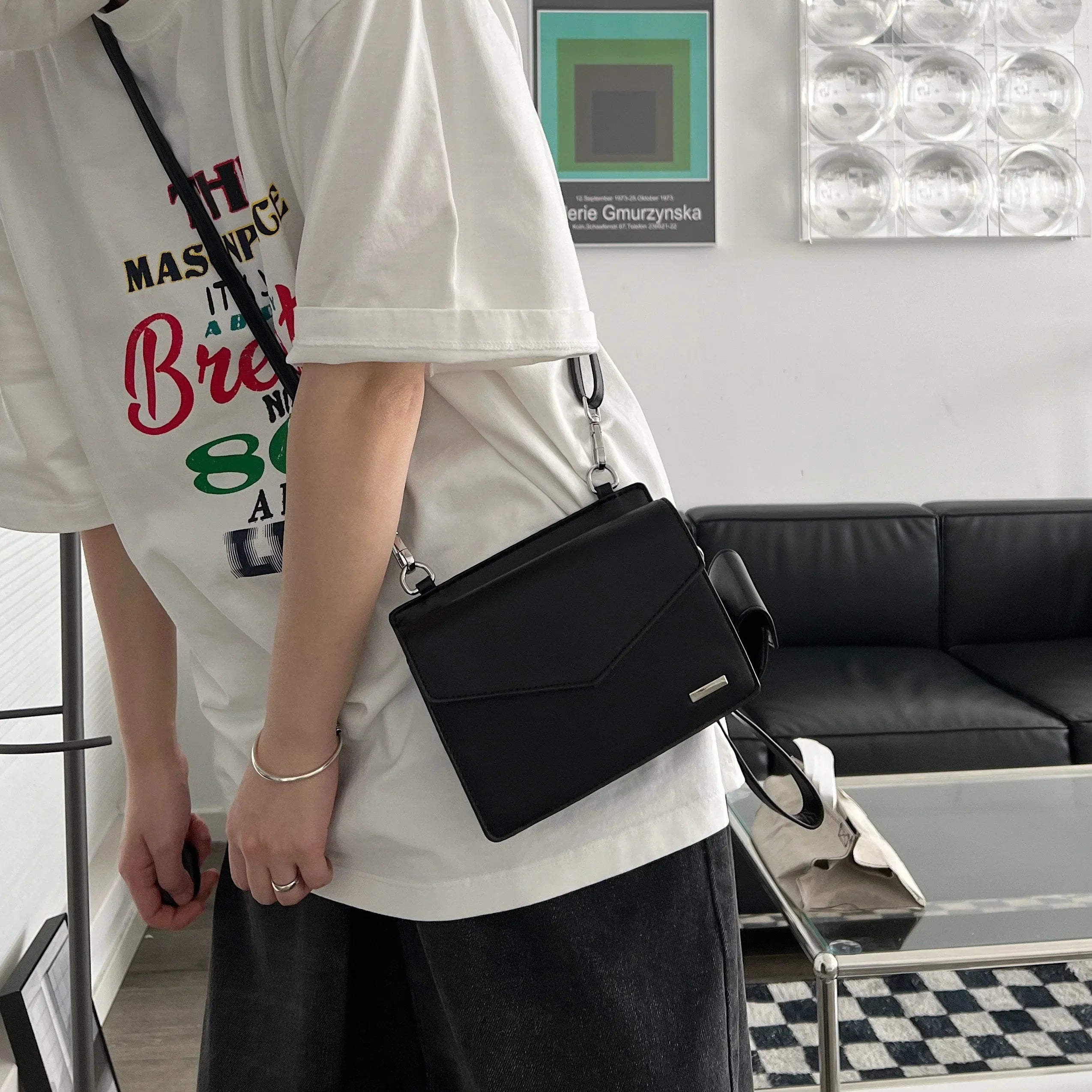 Borsa a tracolla nera in pelle, indossata da una persona con t-shirt bianca e pantaloni scuri, in un salotto moderno. Accessorio moda elegante.