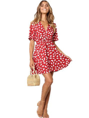 Abito estivo rosso a pois bianchi, maniche corte, scollo a V, indossato da donna sorridente. Accessori: borsa paglia, sandali beige.