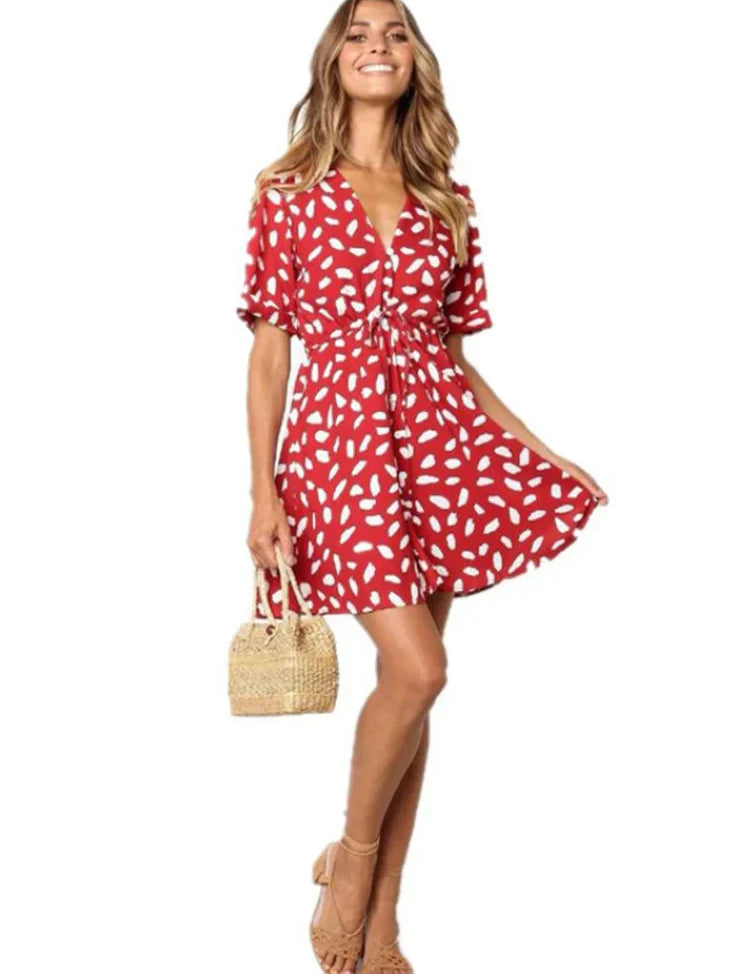 Abito estivo rosso a pois bianchi, maniche corte, scollo a V, indossato da donna sorridente. Accessori: borsa paglia, sandali beige.