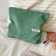Borsa cosmetica verde in tessuto a coste su letto bianco, con cerniera e etichetta "Sozakka". Accessori trucco e cuscino a righe sullo sfondo.
