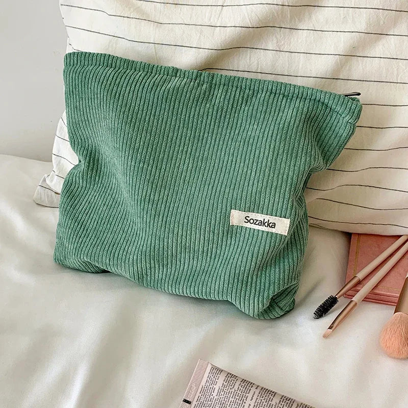 Borsa cosmetica verde in tessuto a coste su letto bianco, con cerniera e etichetta "Sozakka". Accessori trucco e cuscino a righe sullo sfondo.