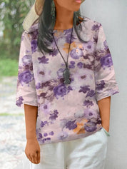 Donna con maglia floreale viola e bianca, maniche a tre quarti, collana nera, cappello di paglia, pantaloni bianchi. Moda casual estiva.