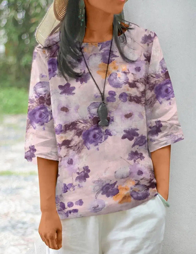 Donna con maglia floreale viola e bianca, maniche a tre quarti, collana nera, cappello di paglia, pantaloni bianchi. Moda casual estiva.