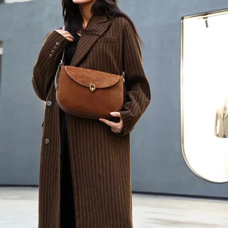 Cappotto lungo a righe marroni con borsa a tracolla in pelle marrone, chiusura a fibbia dorata. Moda autunnale elegante per donna.
