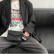 Giacca nera elegante su divano, borsa a tracolla nera in pelle, pantaloni grigi, maglietta bianca con stampa colorata. Moda casual uomo.