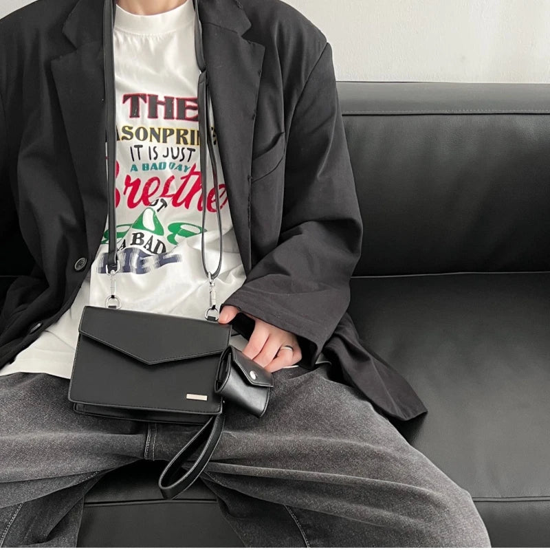 Giacca nera elegante su divano, borsa a tracolla nera in pelle, pantaloni grigi, maglietta bianca con stampa colorata. Moda casual uomo.