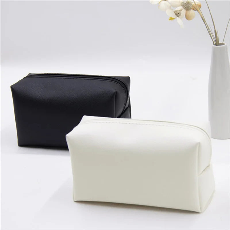 Due astucci in pelle sintetica, uno nero e uno bianco, su sfondo bianco con vaso di fiori. Eleganti e pratici per cosmetici o accessori da viaggio.