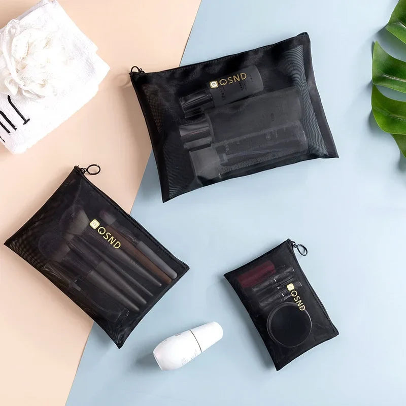 Set di tre pochette trasparenti nere per cosmetici, contenenti trucchi e accessori, su sfondo chiaro con foglia verde e asciugamano bianco.