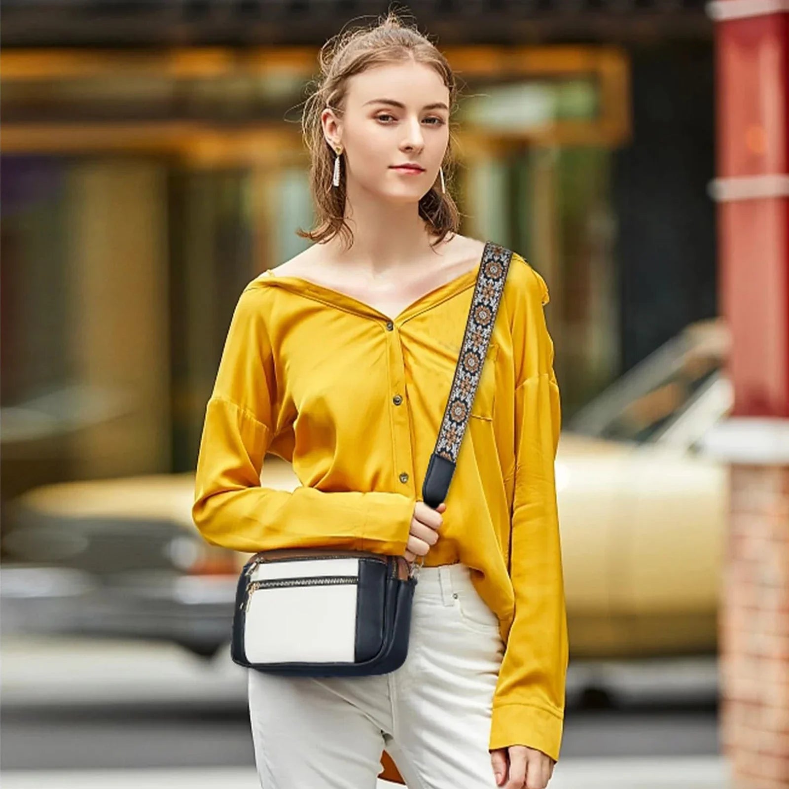 Giovane donna con camicia gialla e borsa a tracolla bianca e nera, sfondo urbano sfocato. Moda femminile, stile casual chic.