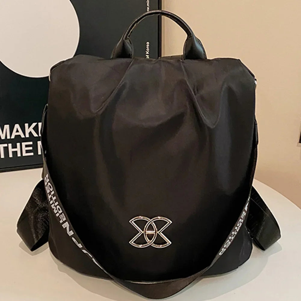 Zaino nero elegante con logo decorativo, manico superiore e tracolla regolabile. Design moderno, ideale per viaggi e uso quotidiano.