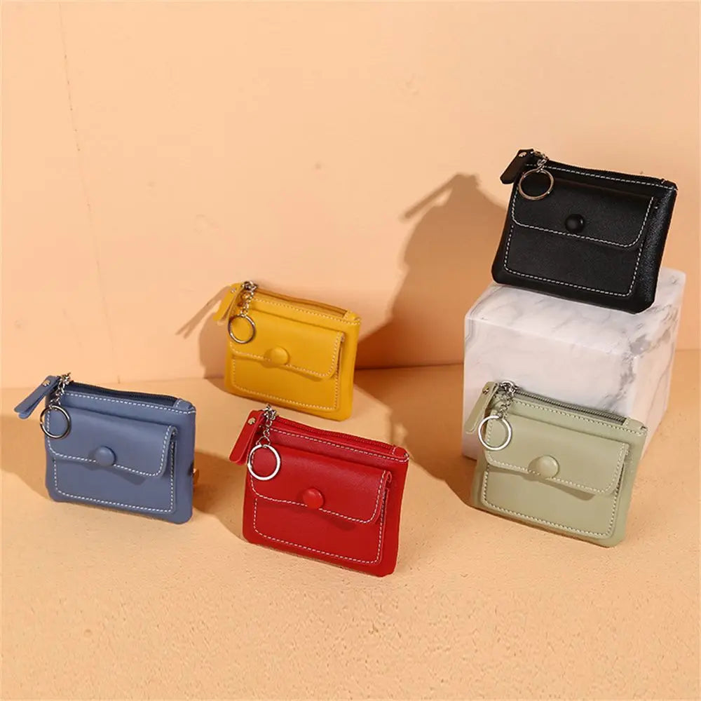 Cinque portamonete in pelle colorata: nero, giallo, blu, rosso e verde, con cerniera e anello portachiavi, su sfondo beige. Accessori moda eleganti.