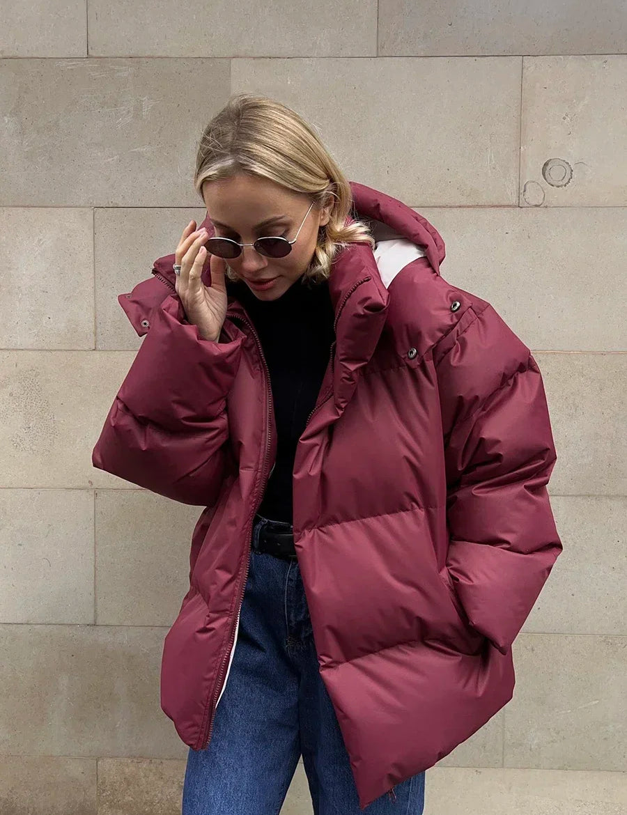 Giacca invernale donna, piumino rosso borgogna, cappuccio, stile casual, moda autunno inverno, occhiali da sole, jeans blu, sfondo muro beige.