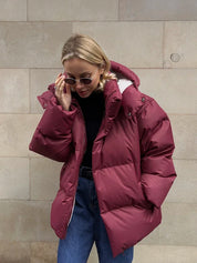 Giacca invernale donna, piumino rosso borgogna, cappuccio, stile casual, moda autunno inverno, occhiali da sole, jeans blu, sfondo muro beige.