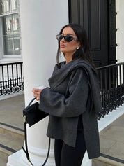 Donna con occhiali da sole, indossa maglione grigio oversize e pantaloni neri, tiene borsa nera. Sfondo urbano elegante con colonne bianche.