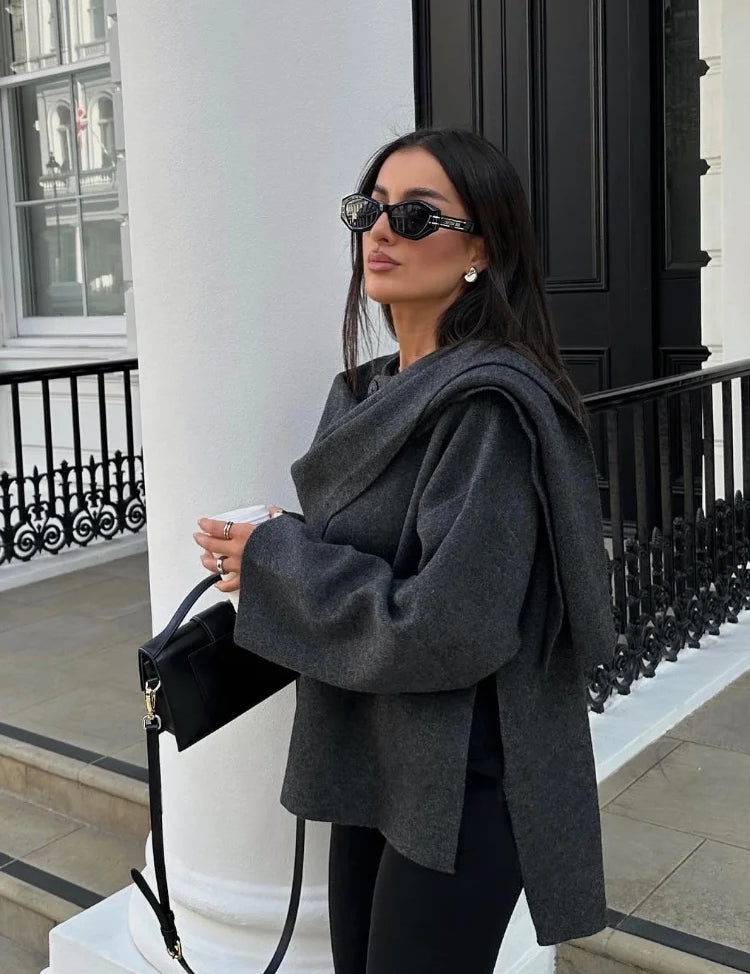 Donna con occhiali da sole, indossa maglione grigio oversize e pantaloni neri, tiene borsa nera. Sfondo urbano elegante con colonne bianche.