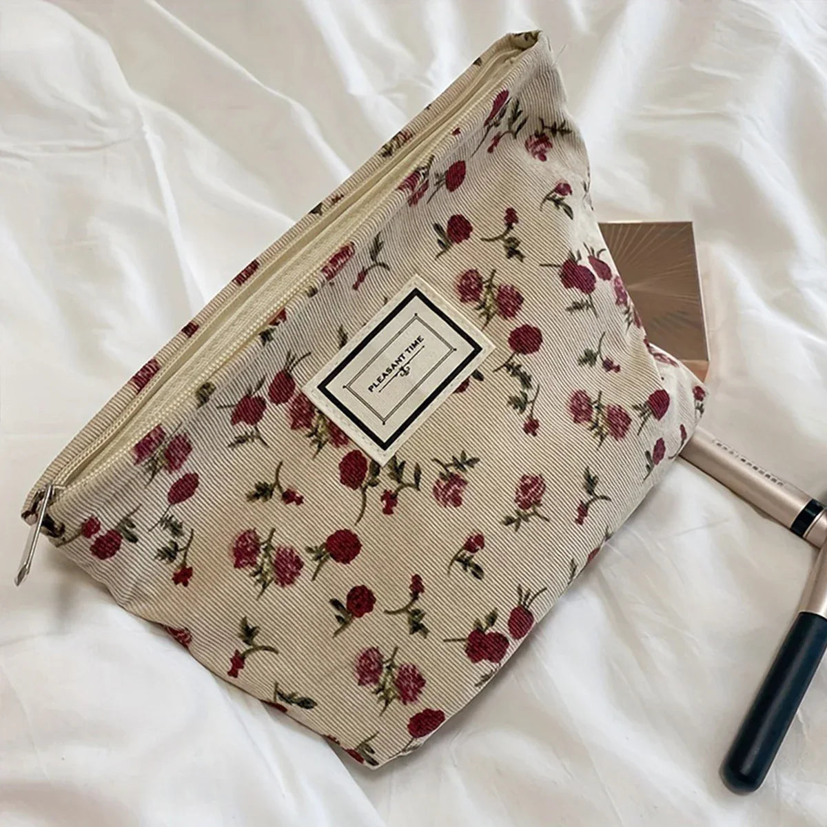 Pochette cosmetica floreale su letto bianco, con cerniera e motivo rose rosse. Accessori trucco accanto. Elegante e pratica per viaggi.