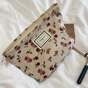 Pochette cosmetica floreale su letto bianco, con cerniera e motivo rose rosse. Accessori trucco accanto. Elegante e pratica per viaggi.