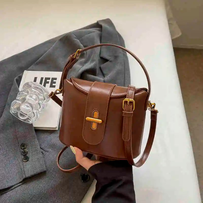 Borsa a tracolla in pelle marrone su giacca grigia, con chiusura dorata e manico regolabile. Accessorio moda elegante per donna.
