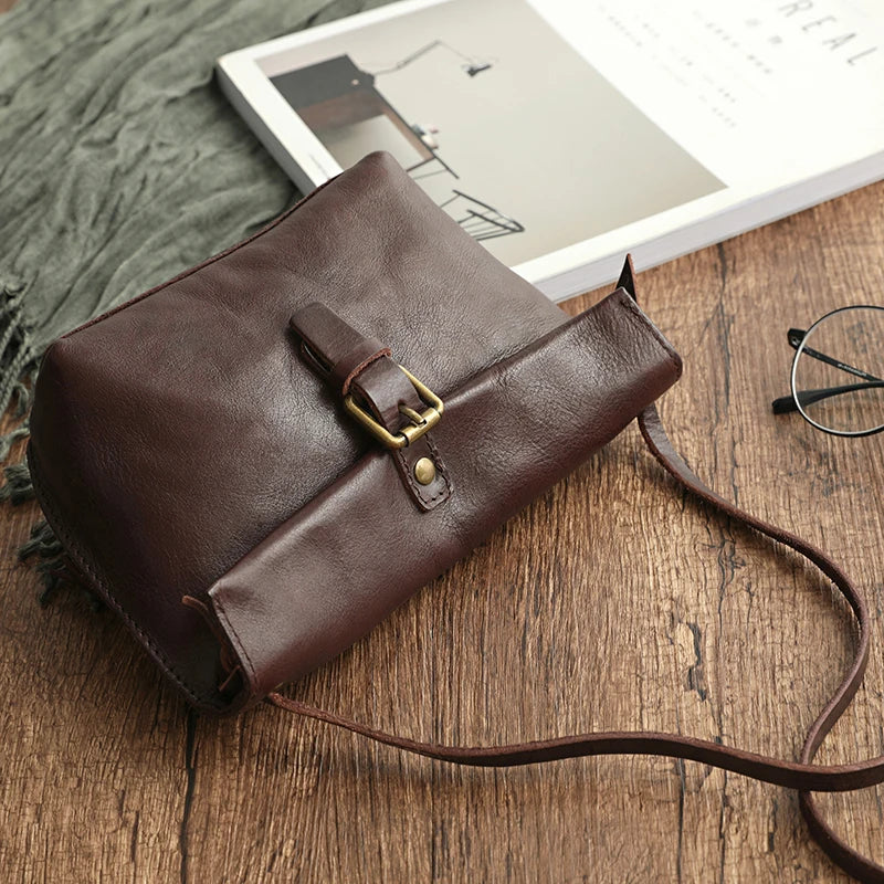 Borsa a tracolla in pelle marrone su tavolo di legno, con occhiali e libro. Design elegante, chiusura a fibbia, accessorio moda donna.