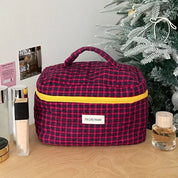 Borsa cosmetica a quadri rossi e blu con cerniera gialla, su tavolo accanto ad albero di Natale e profumi. Ideale per organizzare trucchi e accessori.