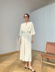Donna in abito bianco elegante, occhiali da sole, sandali, in piedi accanto a sedia moderna. Moda estiva, stile minimalista, interni luminosi.