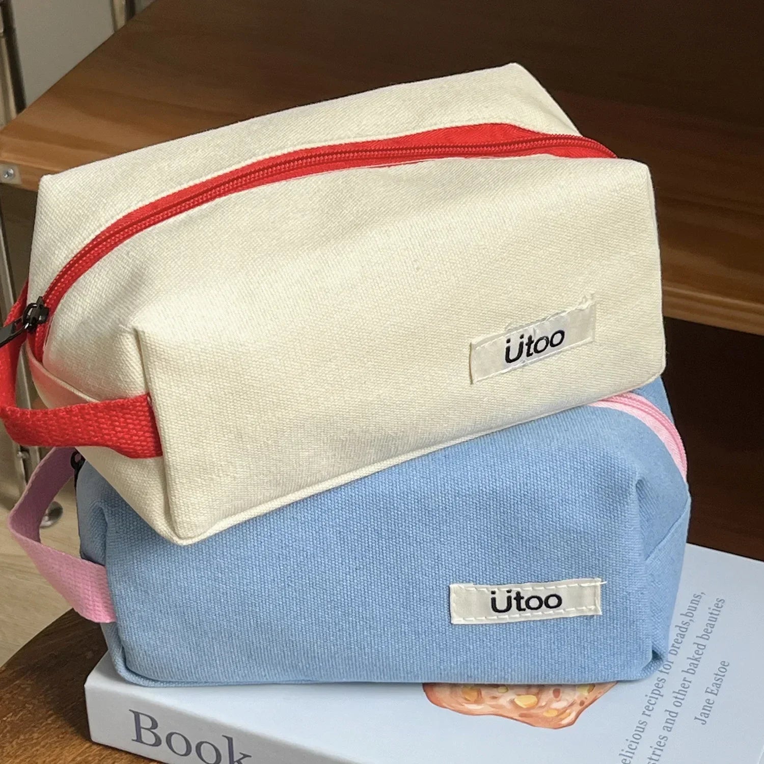 Due astucci in tessuto, uno beige con cerniera rossa e uno azzurro, su un libro. Ideali per scuola o viaggio. Marca Utoo.