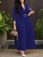 Abito lungo blu elegante con cintura, maniche lunghe e gonna plissettata. Donna con borsa beige e tacchi bianchi. Outfit moda femminile sofisticato.