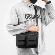 Uomo con giacca grigia e borsa a tracolla nera con tasche multiple. Moda urbana, accessori pratici, stile casual.
