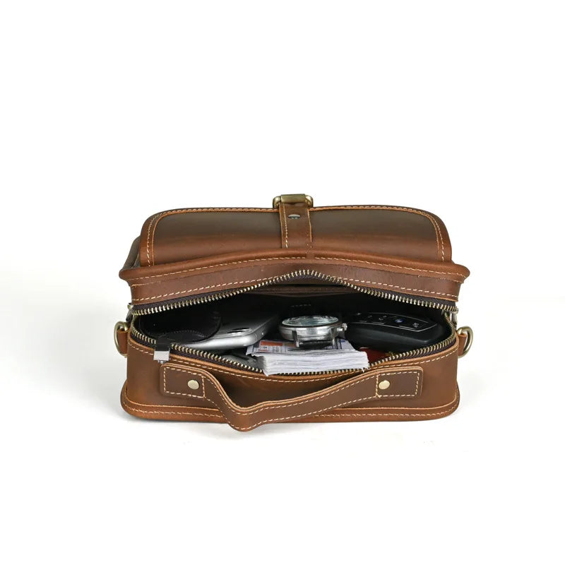 Borsa a tracolla in pelle marrone aperta, con zip, contenente oggetti personali. Design elegante, ideale per uomo e donna. Accessorio moda versatile.