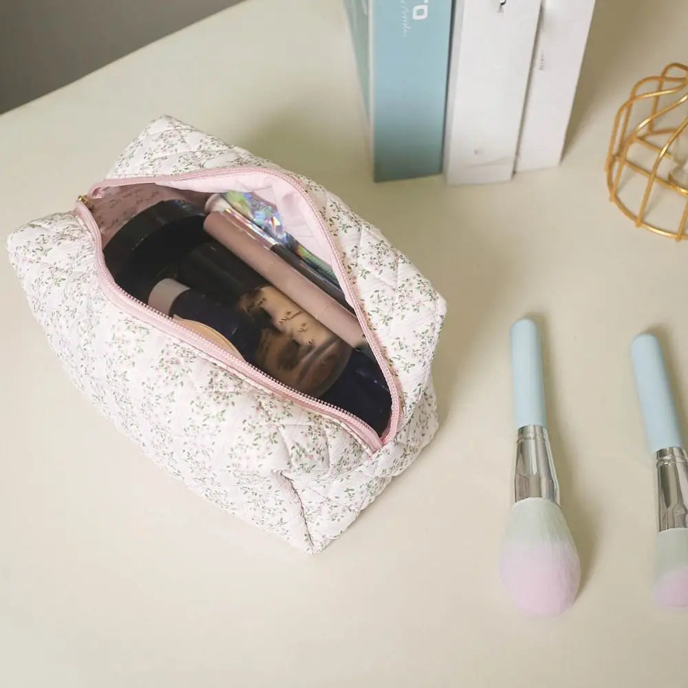 Beauty case floreale aperto su tavolo bianco, contenente trucchi e accessori. Due pennelli da trucco con manico azzurro sono accanto.