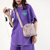 Donna con maglietta viola e borsa a tracolla rosa, tiene cuffie bianche. Moda casual, accessori femminili, stile urbano.
