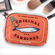Scatola di sardine in olio d'oliva su sfondo bianco, circondata da pennelli e trucco. Design vintage, pesce blu su etichetta rossa.