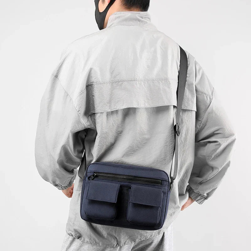 Uomo con giacca grigia indossa borsa a tracolla blu scuro con tasche multiple, design moderno e funzionale, ideale per viaggi e uso quotidiano.