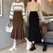 Due donne in gonna a vita alta e maglione, una con stivali bianchi e borsa bianca, l'altra con stivali neri e borsa marrone, in un salotto moderno.