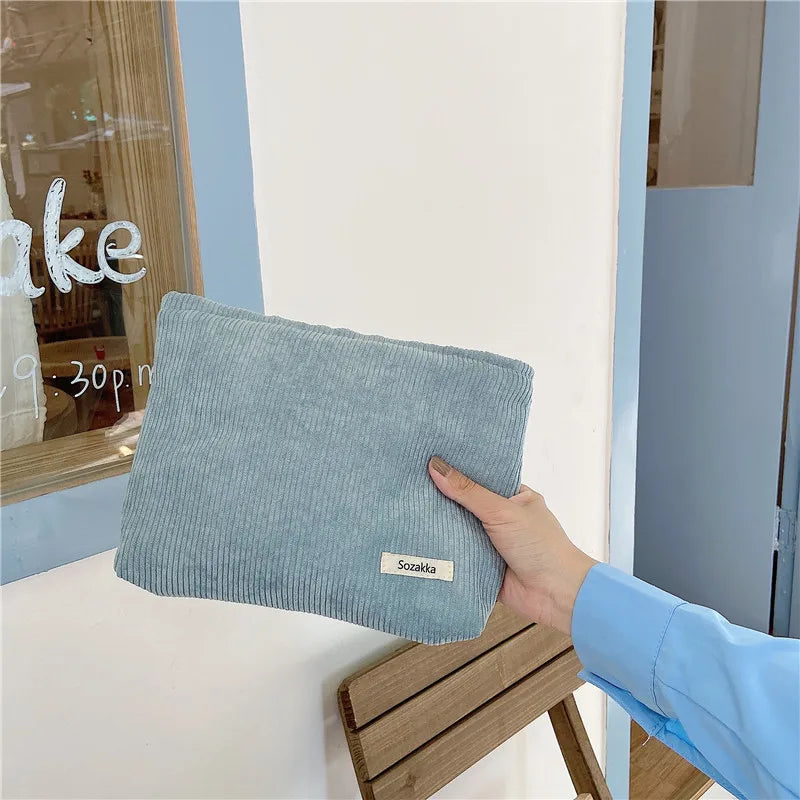 Mano che tiene una borsa a tracolla in velluto a coste azzurro, marchio Sozaka, davanti a una porta blu. Accessorio moda elegante e moderno.