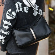 Borsa a tracolla in pelle nera con striscia centrale grigia, indossata su felpa con cappuccio. Accessorio moda uomo, stile casual ed elegante.