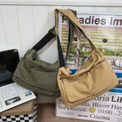 Due borse a tracolla in tela, una verde oliva e una beige, appoggiate su una scrivania accanto a un laptop. Design casual e spazioso.