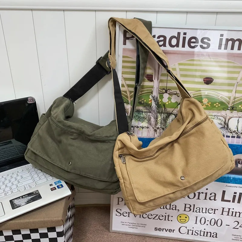 Due borse a tracolla in tela, una verde oliva e una beige, appoggiate su una scrivania accanto a un laptop. Design casual e spazioso.
