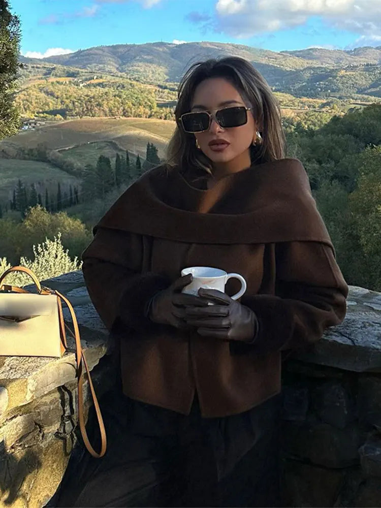 Donna con occhiali da sole e cappotto marrone, tiene una tazza di caffè su una terrazza panoramica in campagna. Paesaggio collinare autunnale sullo sfondo.